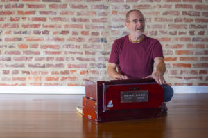 Hari-kirtana das sitting on wood floor with a harmonium