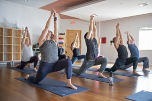 Brightly lit yoga class of students performing a variation of Warrior 1 pose