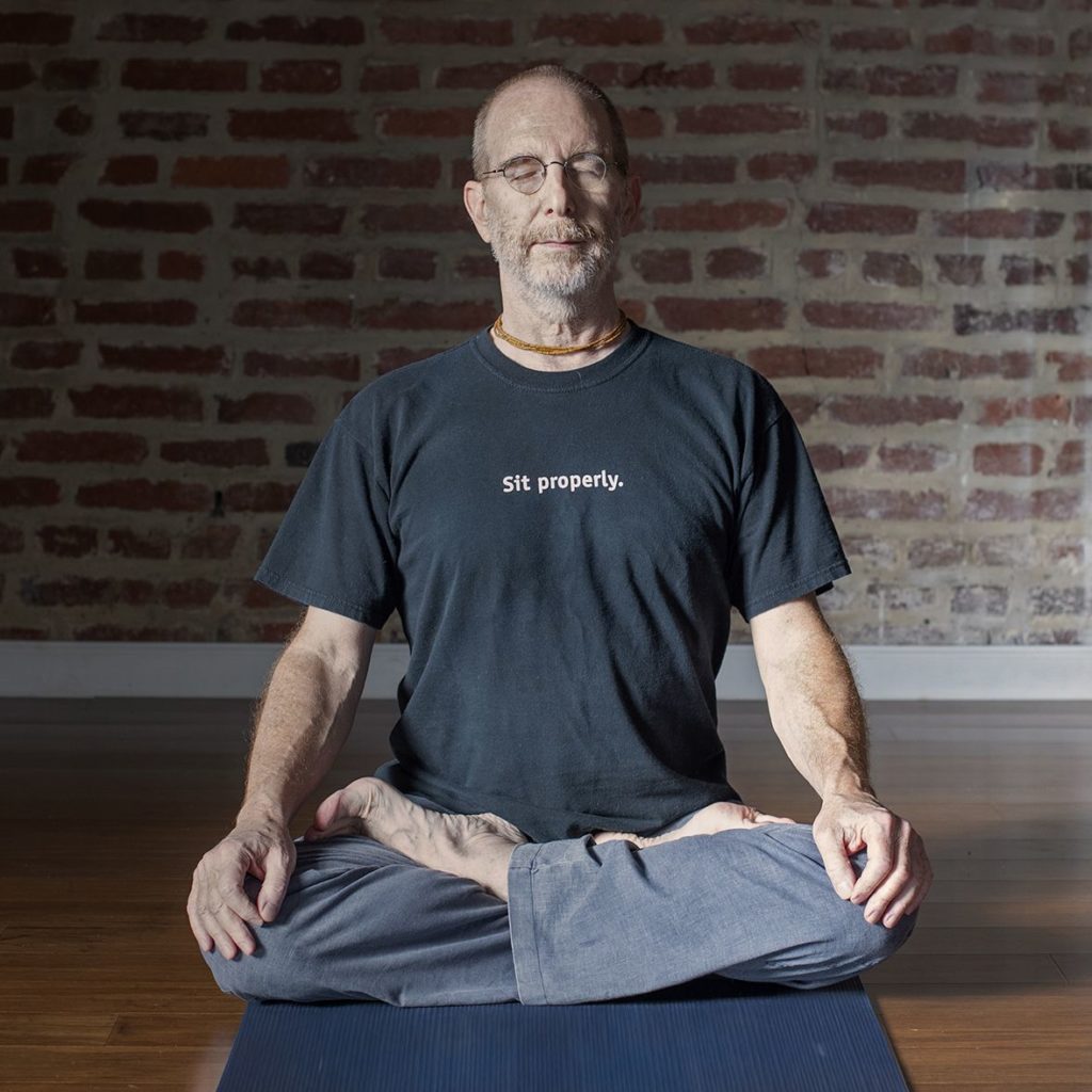 Hari-kirtana das sitting on wood floor meditating in front of a brick wall