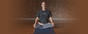 Hari-kirtana das sitting on wood floor meditating in front of a brick wall