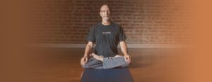 Hari-kirtana das sitting on wood floor meditating in front of a brick wall